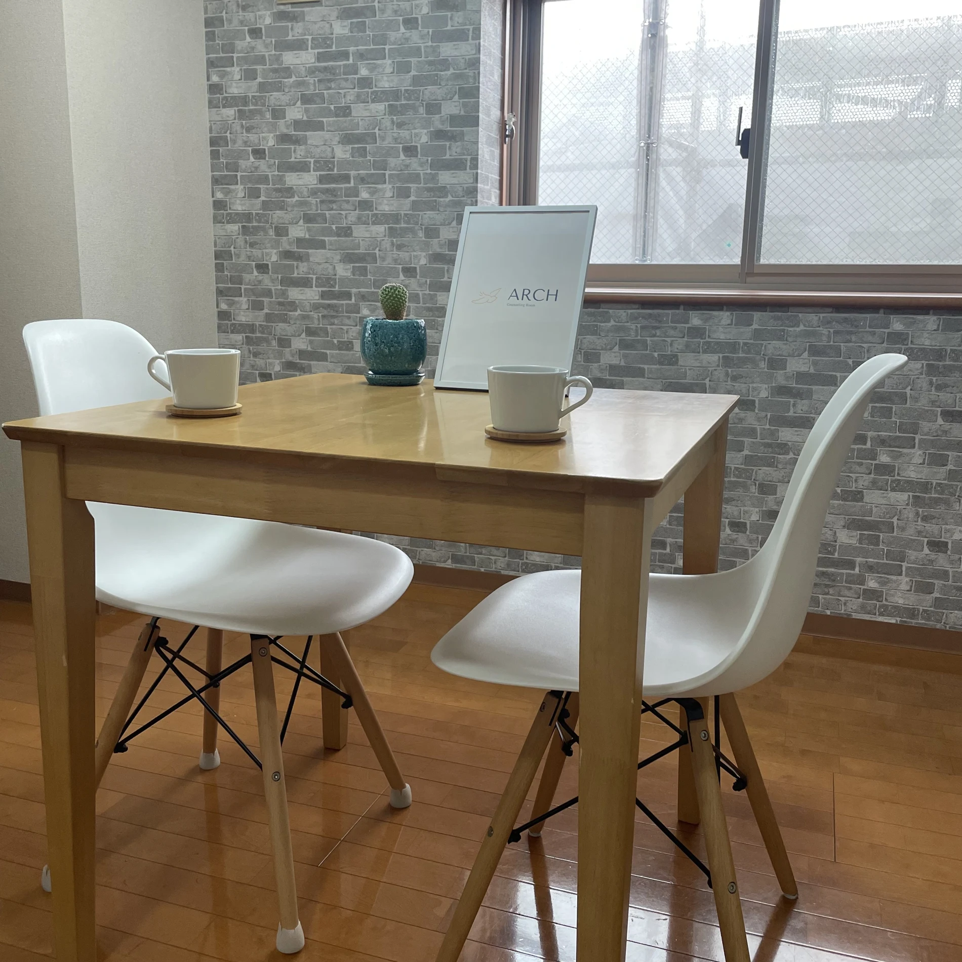 窓から光が入るARCHのお部屋。木のテーブルと二脚の椅子、二つのコーヒーカップ。